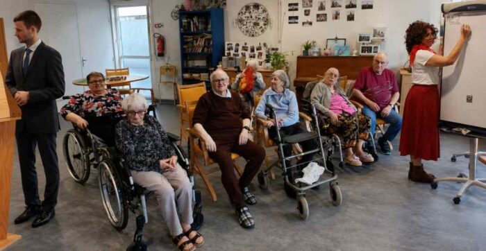 un groupe de résidents dans un Ehpad sont attentifs face à une jeune femme qui écrit sur un tableau, un homme en costume se tient à côté